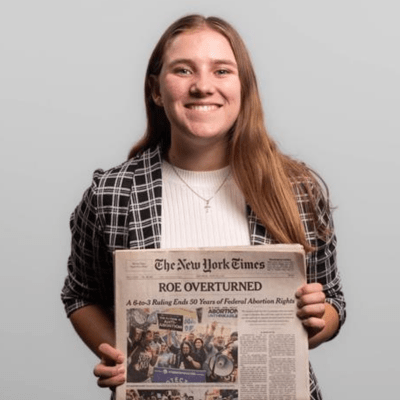 A woman holds a New York Times newspaper and smiles at the camera.