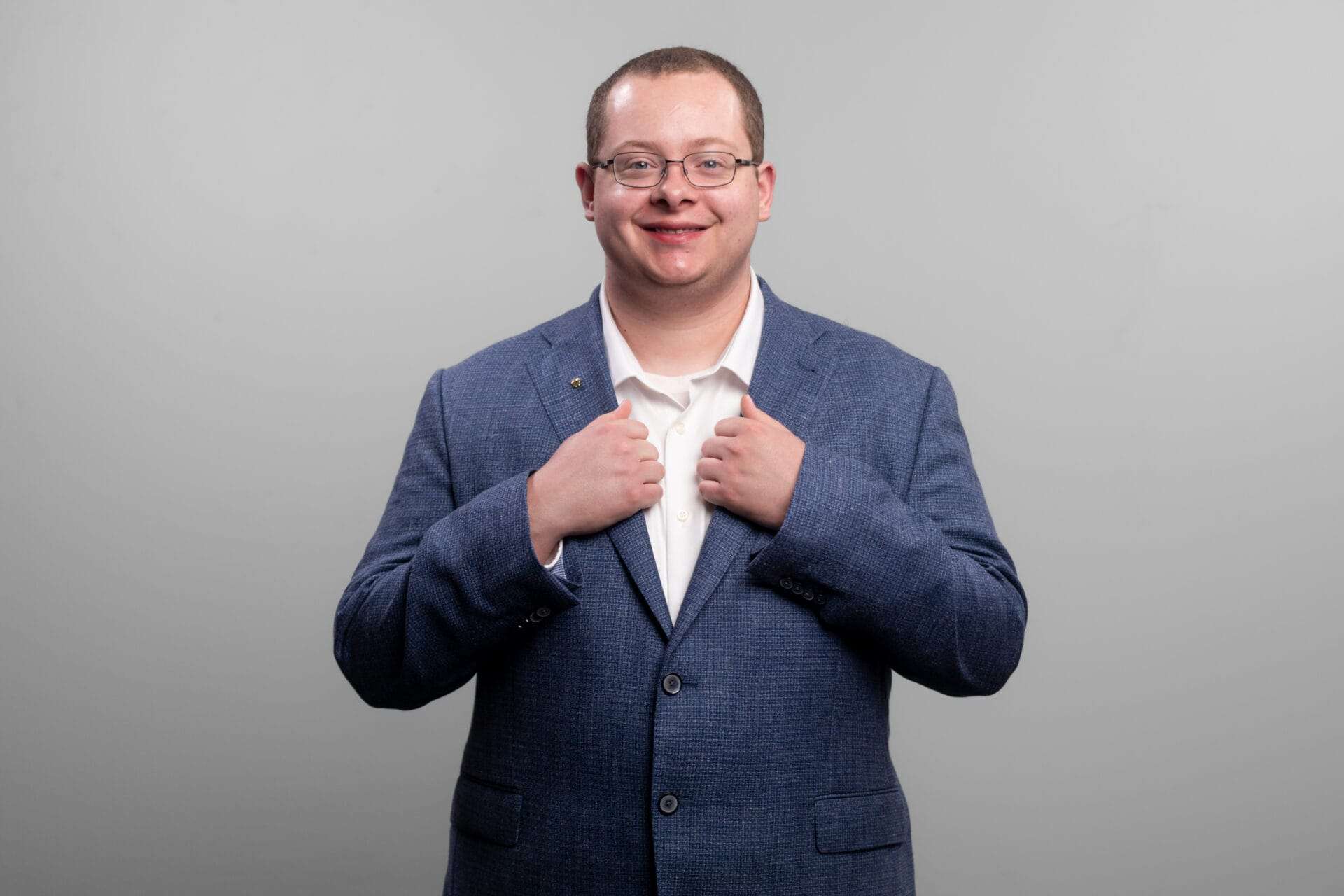 A man with glasses grabs the opening of his suit on either side with his hands as he smiles at the camera.