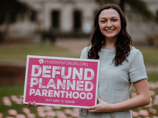 A woman smiles and holds a sign that reads, "DEFUND PLANNED PARENTHOOD."
