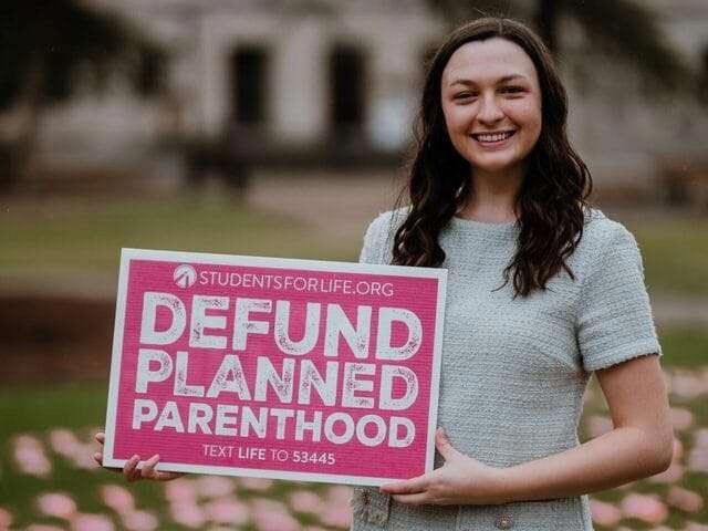 A woman smiles and holds a sign that reads, "DEFUND PLANNED PARENTHOOD."