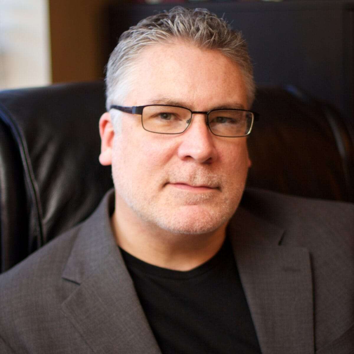 A man wearing a black shirt, suit jacket with no tie, and glasses