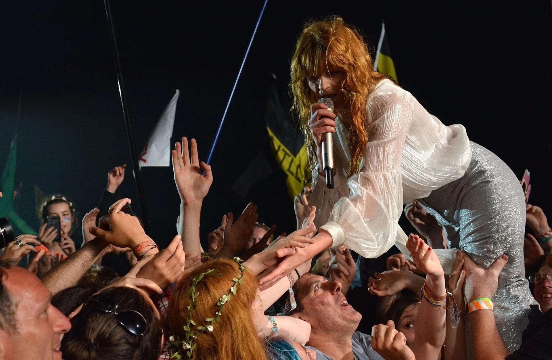 Florence Welch sings into a microphone and reaches out into the crowd. The crowd's hands are in the air.