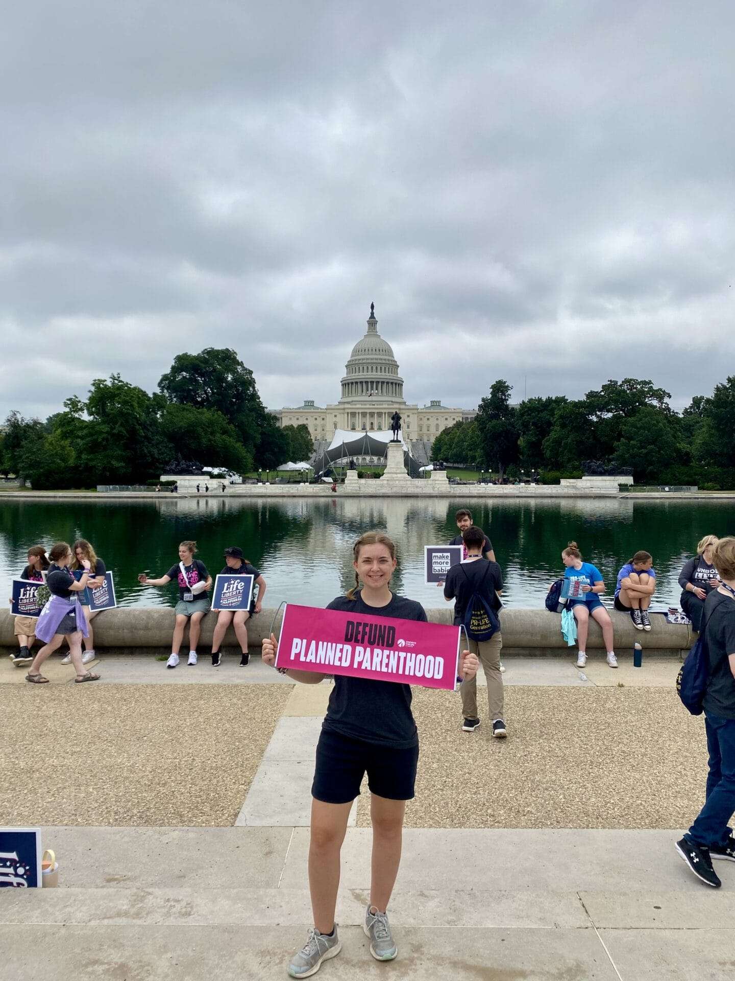 A woman holds a sign that advocates for the defunding of Planned Parenthood. The capitol building can be seen in the background.